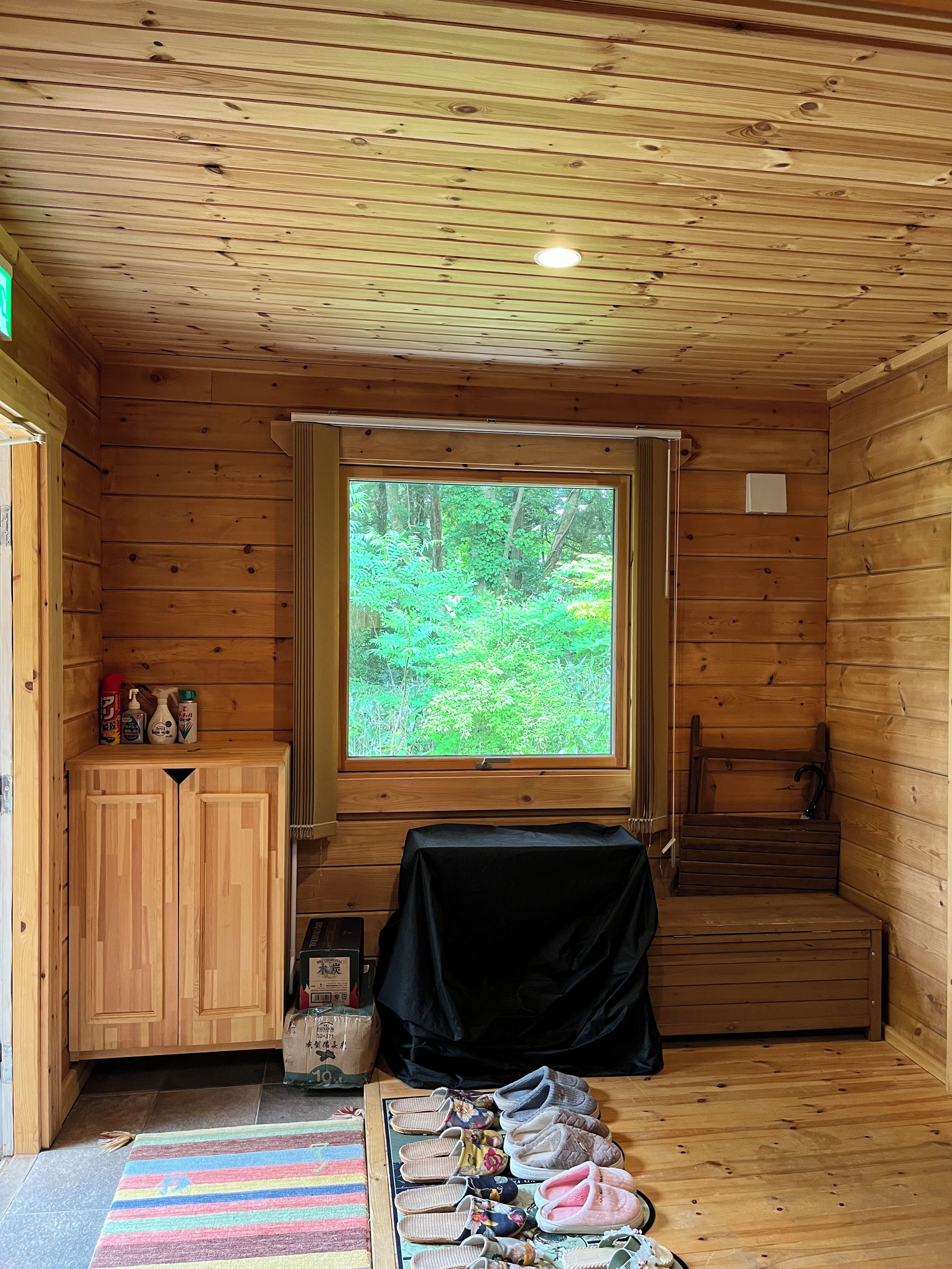 A wooden cabin interior with a large window, a wooden cabinet, a covered table, and a pair of steps leading to a loft area.