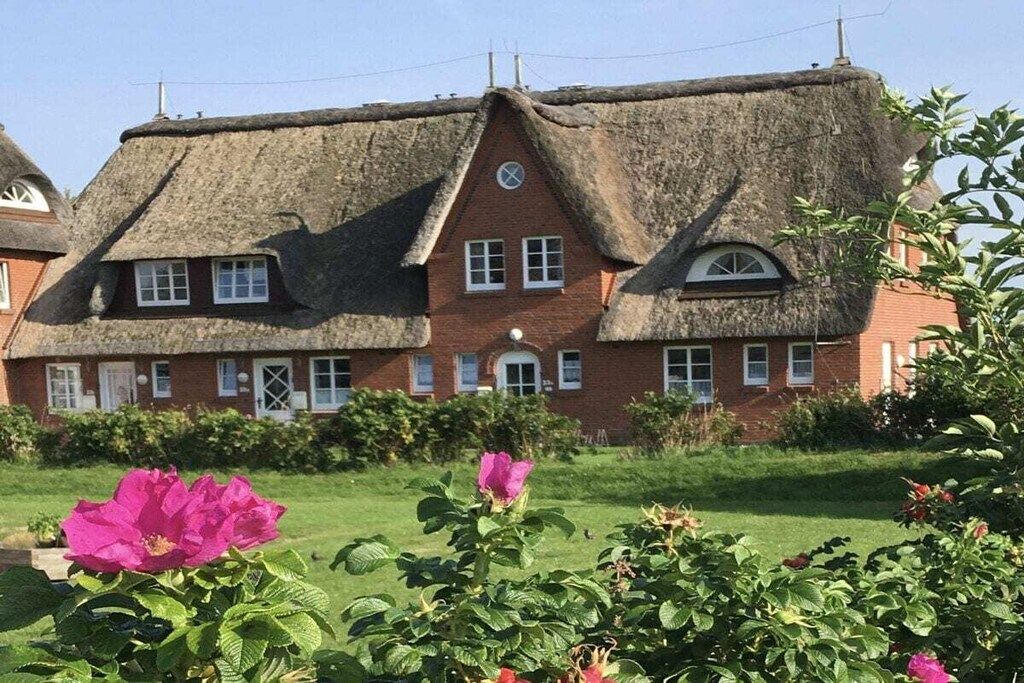 Ein traditionelles Reetdachhaus mit Ziegelverkleidung, mehreren Fenstern und einem gepflegten Garten mit blühenden Pflanzen.