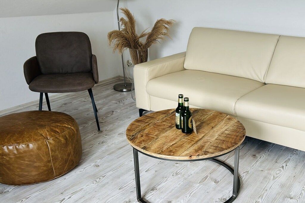 Ein Wohnzimmer mit einem braunen Ledersitzhocker, einem braunen Sessel, einem runden Holztisch mit zwei Flaschen und einem cremefarbenen Sofa.