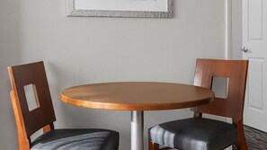 Una mesa redonda de madera con dos sillas, cada una con un corte cuadrado en el respaldo, en una habitación con una pared lisa y una puerta.