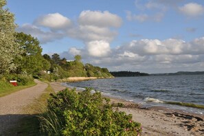 A gravel path leading to a beach with a view of the water and distant hills. - Holiday home with 2 bedrooms (Brodersby)