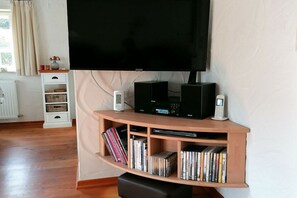 A living room with a flat-screen TV mounted on the wall, a wooden entertainment unit with a DVD player, and a white shelf with decorative items. - Holiday home with 2 bedrooms (Brodersby)