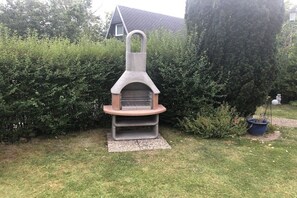 An outdoor stone-built barbecue grill with a chimney, surrounded by a well-maintained hedge and a potted plant. - Holiday home with 2 bedrooms (Friedrichskoog)