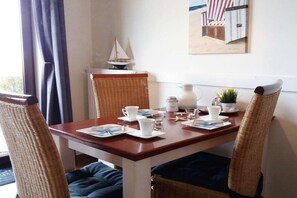 Mesa de comedor dispuesta con platos blancos, tazas y una jarrón, rodeada de sillas de mimbre con cojines.