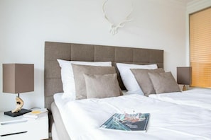 A neatly made bed with white bedding and two pillows, a bedside table with a lamp and a book, and a wall-mounted deer skull decoration. - Holiday apartment with 2 bedrooms (Zingst)