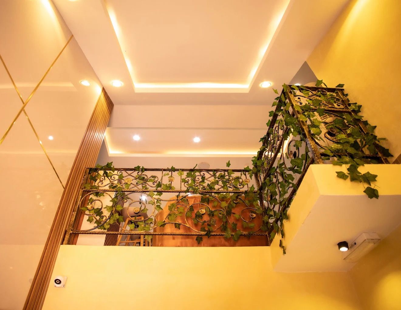Uma escadaria com corrimão decorado com trepadeiras verdes, teto com iluminação embutida e uma câmera de segurança fixada na parede.