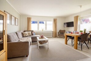 A living room with a sofa, a coffee table, a dining table with chairs, a TV mounted on the wall, and a window with curtains. - Holiday apartment Luzie (Büsum)