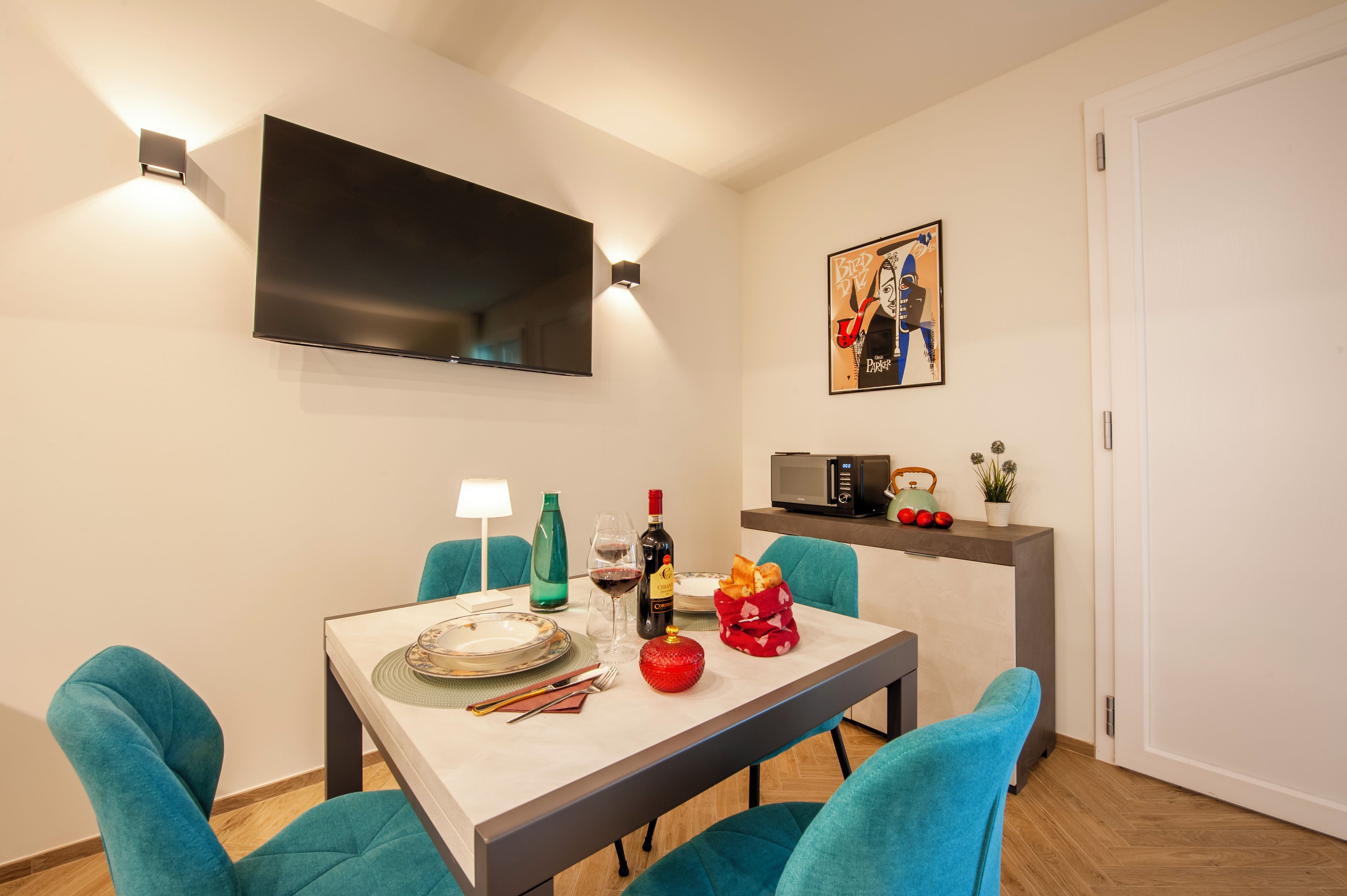 Un comedor con una mesa dispuesta para dos, un televisor de pantalla plana montado en la pared y una obra de arte enmarcada sobre la mesa.