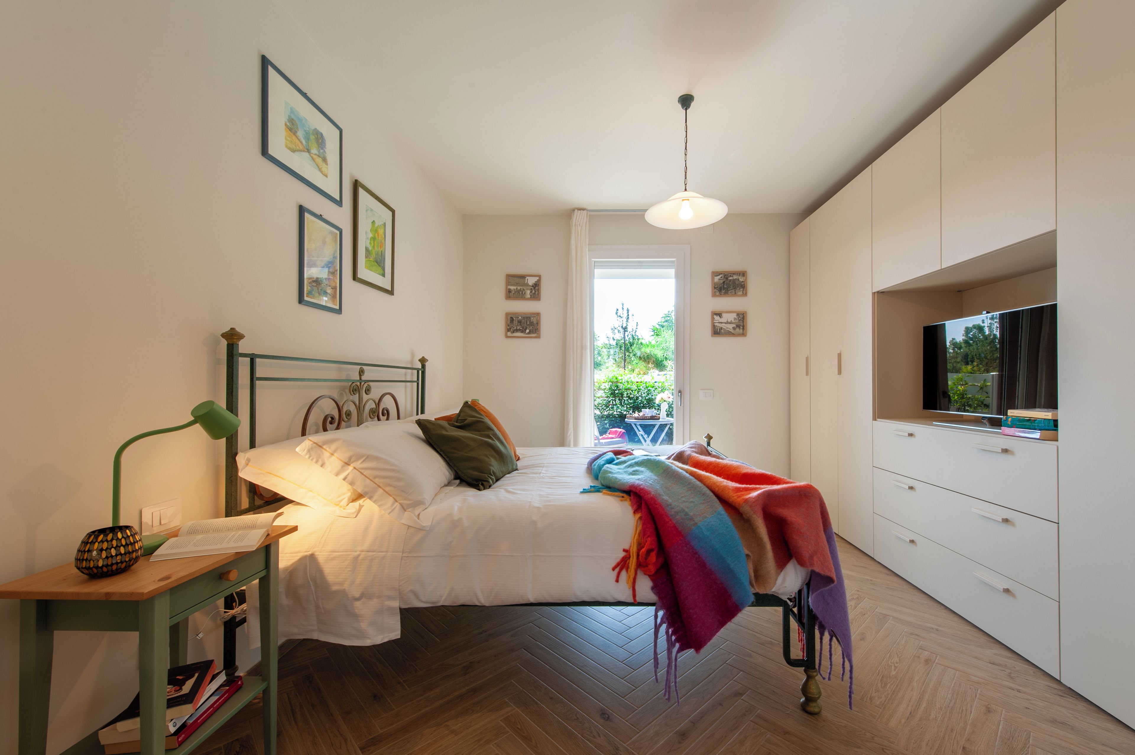 Ein Schlafzimmer mit einem Bett, einem Schreibtisch mit Lampe, einem Fernseher und einem Fenster mit Blick auf Grünflächen.