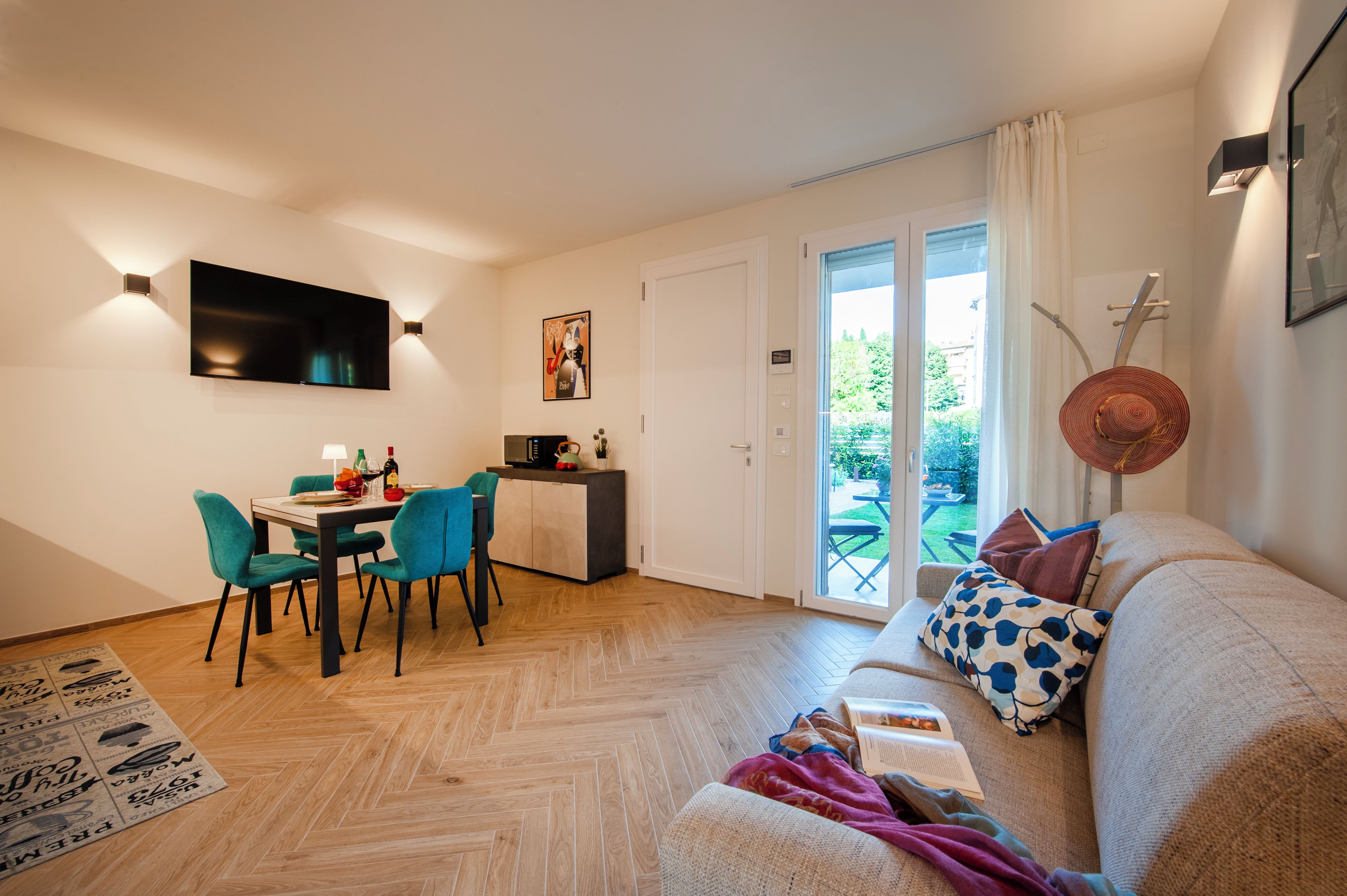 Una sala de estar moderna con un sofá, una mesa de comedor con sillas, un televisor montado en la pared y un ventanal corredero que da a un espacio exterior.