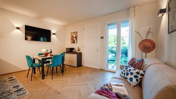 Una sala de estar moderna con un sofá, una mesa de comedor con sillas, un televisor montado en la pared y un ventanal corredero que da a un espacio exterior.