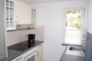 A compact kitchen with white cabinetry, a black countertop, a coffee maker, and a window with a curtain. - Bungalow in Koserow with barbecue (Koserow)