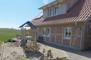 A two-story house with a red-tiled roof, a wooden play structure, and a paved patio area.