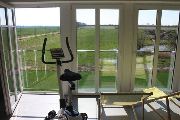 A room with a stationary bike, a chair, and large windows offering a view of a green field.