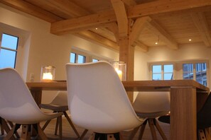 A dining area with a wooden table and chairs, large windows, and a high ceiling with exposed beams.