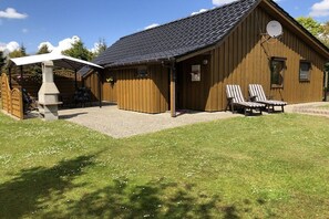 A wooden building with a sloped roof, a covered outdoor seating area, and two lounge chairs. - Holiday home with 2 bedrooms (Börm\/nahe Kropp\/bei Schleswig)