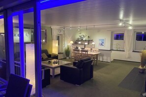 Ein modernes Wohnzimmer mit Glastrennwand, einem Essbereich mit Tisch und Stühlen sowie einer Küche mit Herd und Schränken.