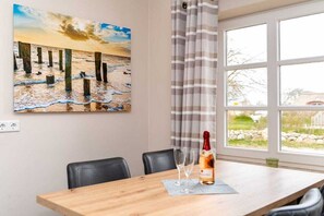 A dining area with a wooden table, two chairs, a bottle of champagne, and two glasses. A painting of a beach scene is on the wall. - Holiday apartment with 2 bedrooms (Oevenum)
