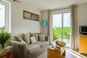 A living room with a sofa, coffee table, and a television. There is a window with curtains, a lamp, and a painting on the wall. - Breton Alcove Stay for Six (Cancale)