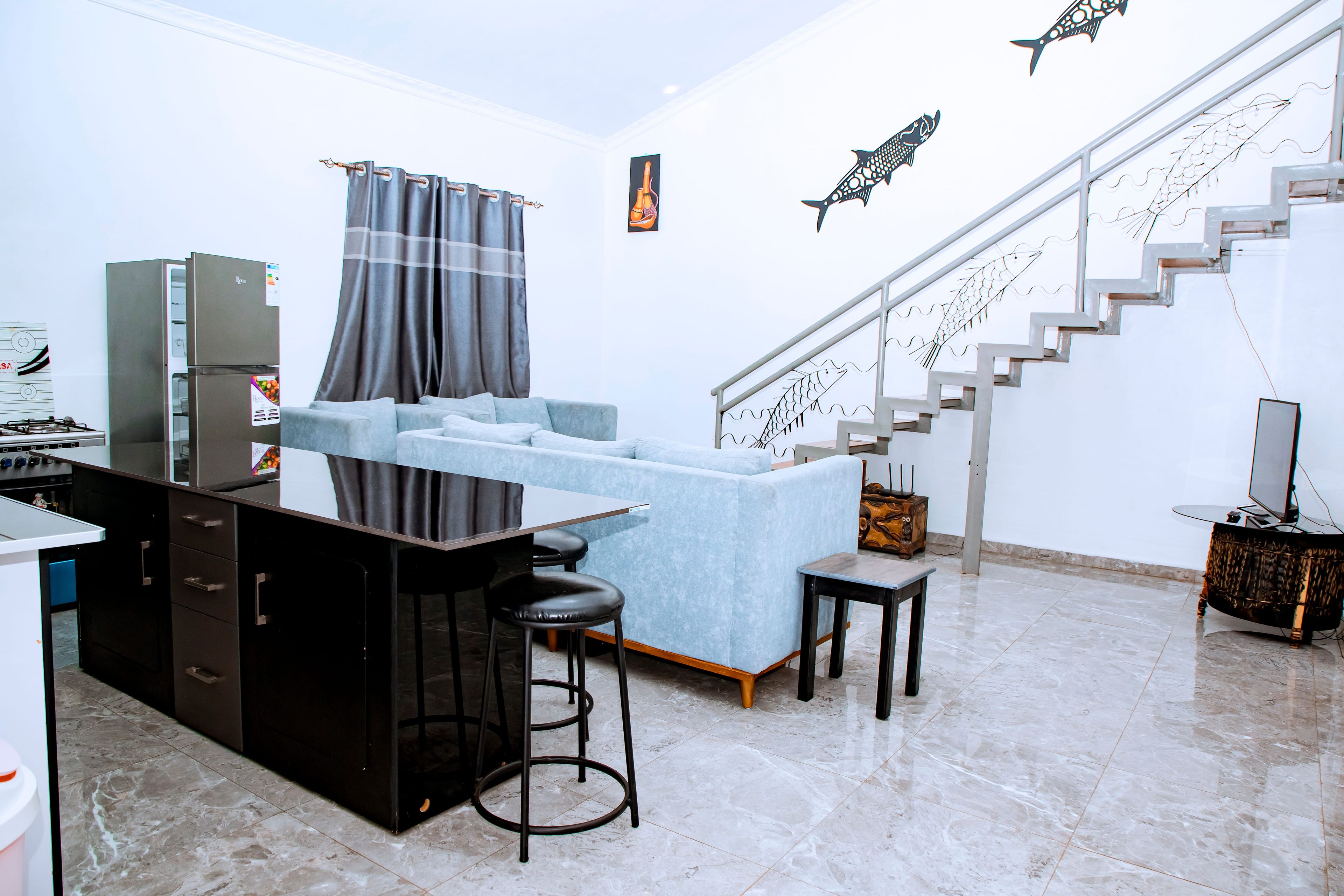 A modern kitchen with a refrigerator, a dining area with a sofa and a small table, and a staircase with decorative fish railings.