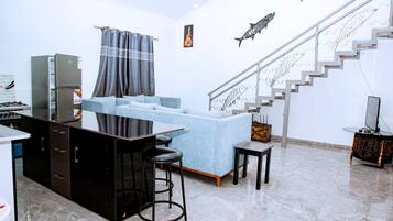 A modern kitchen with a refrigerator, a dining area with a sofa and a small table, and a staircase with decorative fish railings.