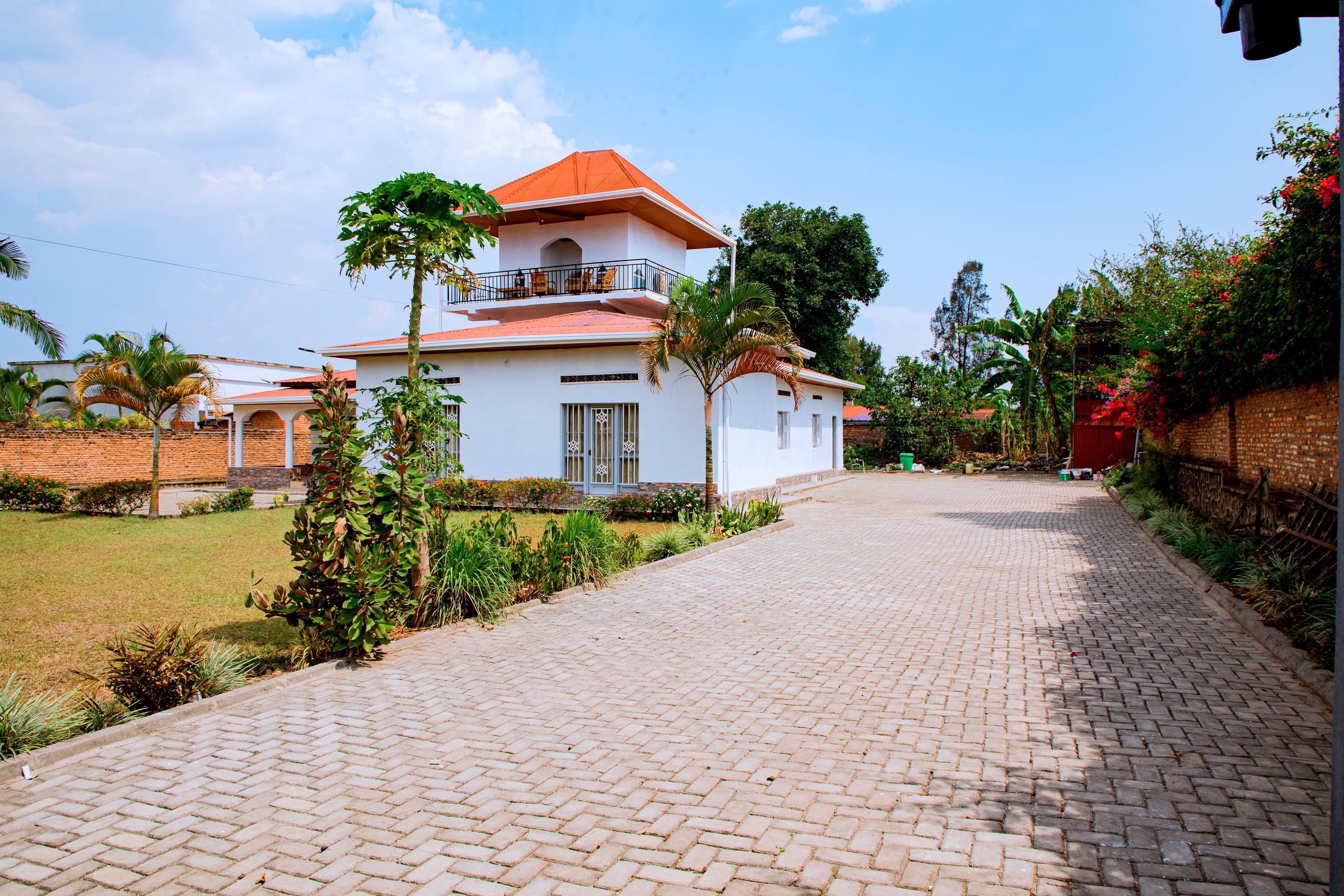 Uma casa de dois andares com telhado vermelho, varanda com móveis e entrada com piso pavimentado.
