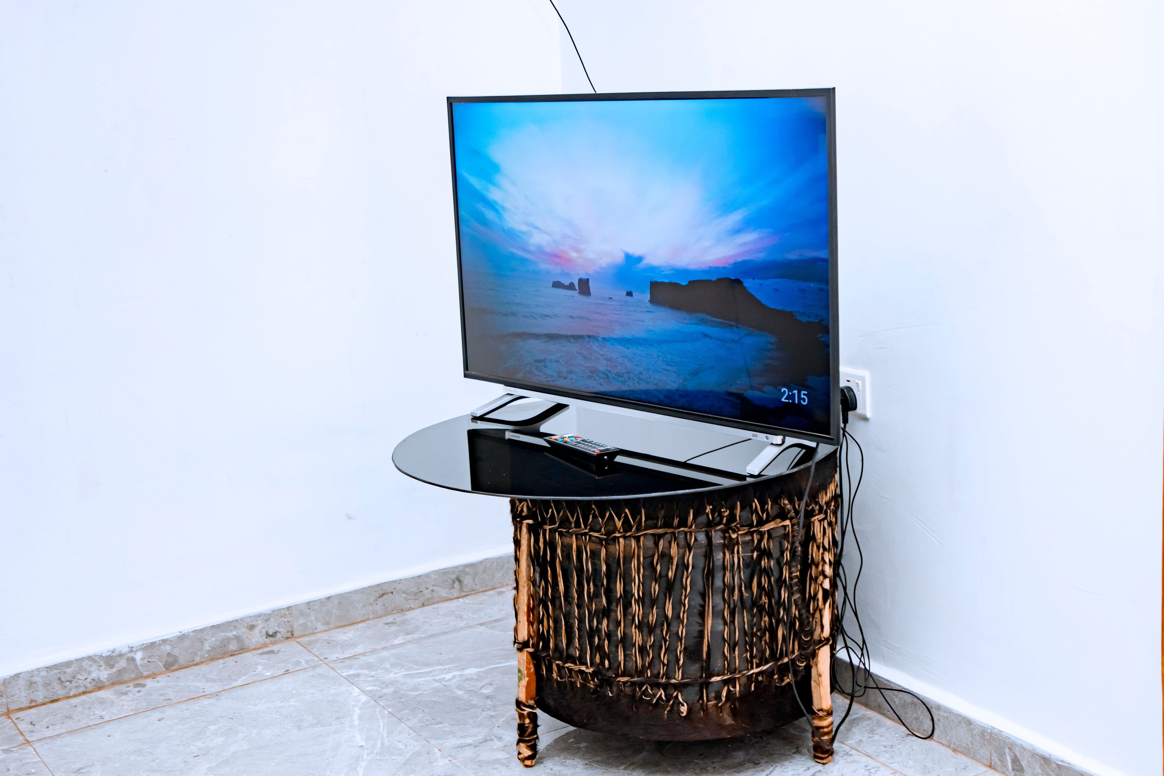 Un televisor de pantalla plana sobre una mesa redonda y trenzada, frente a una pared blanca.