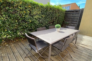 An outdoor patio with a metal table and wicker chairs, surrounded by a wooden deck and a hedge. - Provencal Family Getaway for Six (L'Isle-sur-la-Sorgue)