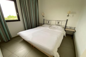 A single bed with a metal frame and a white mattress in a room with a tiled floor and a window with curtains. - Provencal Family Getaway for Six (L'Isle-sur-la-Sorgue)