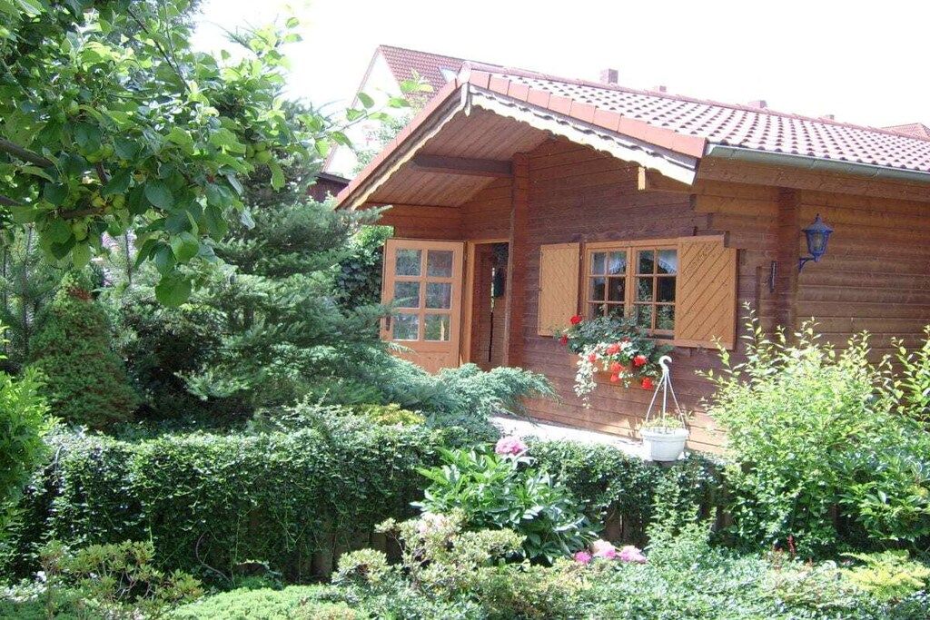A wooden house with a sloped roof, a porch with potted plants, and a garden with various shrubs and flowers.