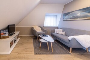 A modern loft-style living room with a sloped ceiling, a grey sofa, a small coffee table, and a television on a white cabinet. - Hein Godenwind - Romeo (St. Peter-Ording)