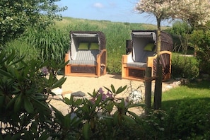 Two wicker lounge chairs with cushions, positioned on a sandy surface surrounded by greenery. - Hein Godenwind - Romeo (St. Peter-Ording)