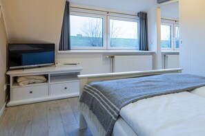 A bedroom with a bed, a television on a stand, a window with curtains, and a radiator. - Hein Godenwind - Romeo (St. Peter-Ording)
