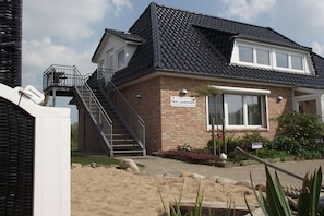 A two-story house with a brick facade, a dark tiled roof, and a staircase leading to a balcony. - Hein Godenwind - Romeo (St. Peter-Ording)