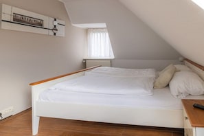 A bedroom with a sloped ceiling, a bed with white bedding, a window with curtains, and a radiator. - Hein Godenwind - Julia (Sankt Peter-Ording)