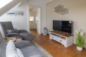 A living room with a sofa, a television on a wooden stand, and a potted plant. - Hein Godenwind - Julia (Sankt Peter-Ording)