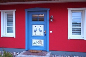 A red building with a blue door featuring a bird and plant design, a small porch with a doormat, and a wall-mounted light fixture. - Boddenhus "Crane Rest" (Born am Darss)