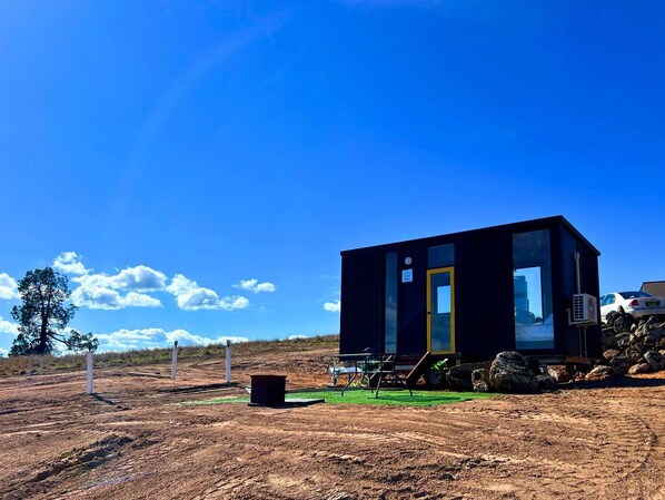 A small, dark-colored cabin with a single visible door and windows, situated in a dry, open landscape with a clear sky.