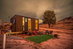 Une cabane moderne dotée d’une grande fenêtre, d’un petit porche et d’un arbre en arrière-plan.