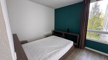 A bedroom with a bed, a window with curtains, a small table, and a wall unit.