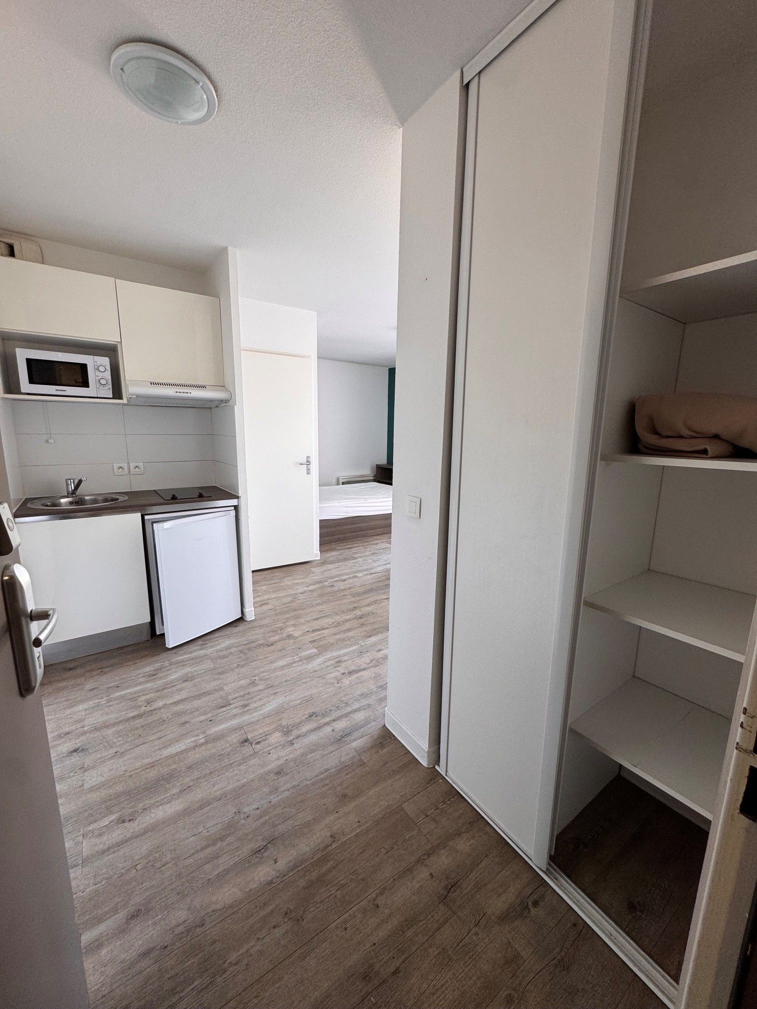 A compact kitchen with a microwave, sink, and a small refrigerator. There is a wooden floor and a white door leading to another room.