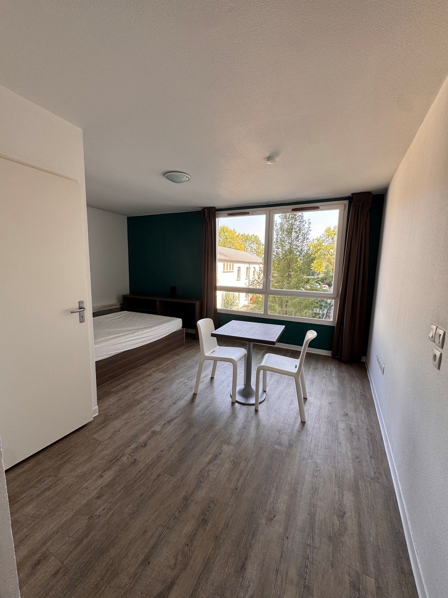 Een kleine kamer met een bed, een tafel en twee stoelen. Er is een raam met gordijnen, en de vloer is van hout.