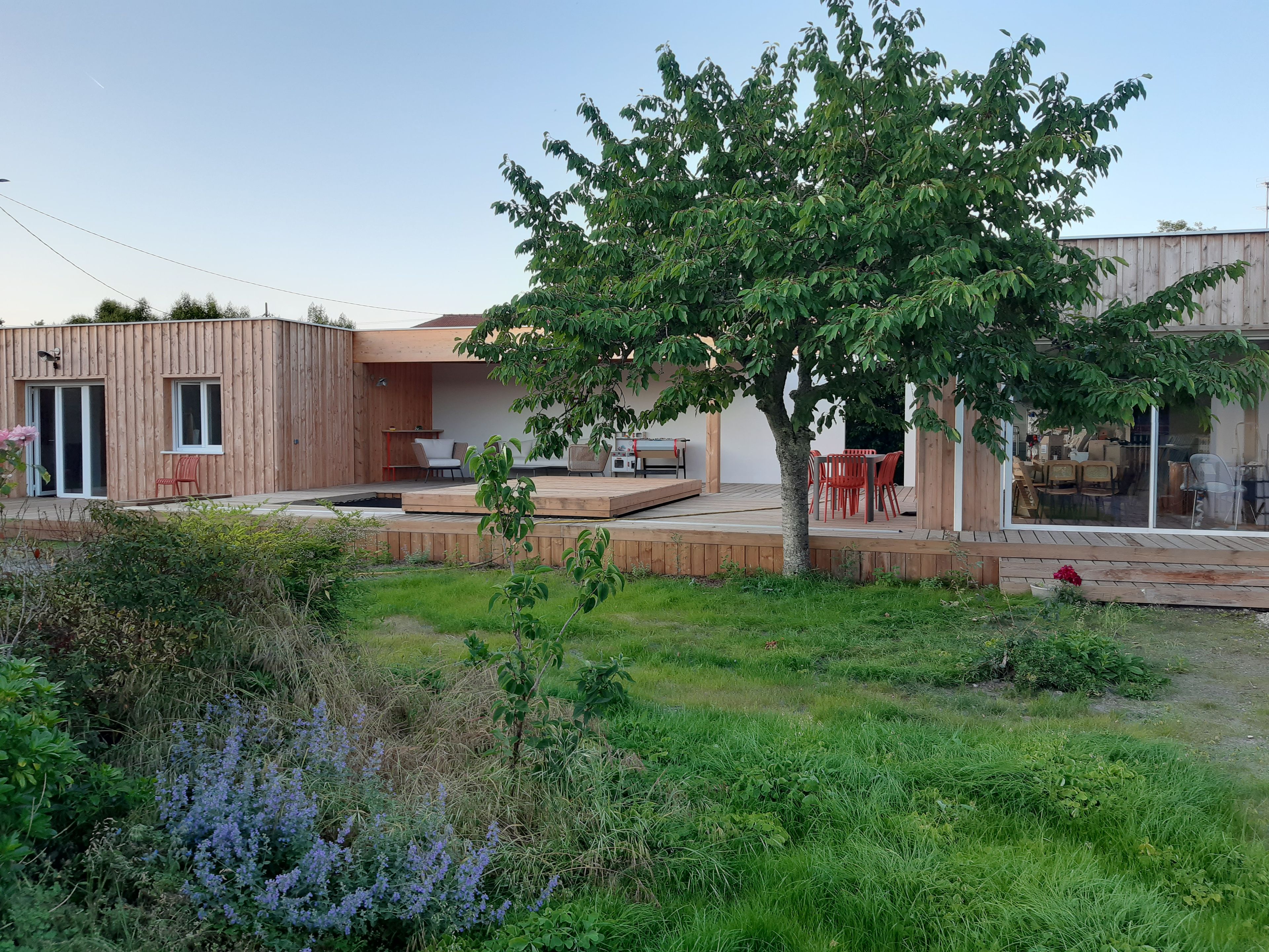 A modern wooden house with a patio area, a dining set, and a large tree.
