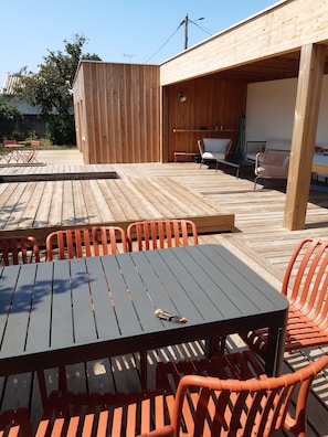 An outdoor wooden deck with tables and chairs, a covered seating area, and a building with a wooden facade. - Spacious, fully equipped house surrounded by greenery, with a pool and a wood stove—perfect for families! (Cestas)