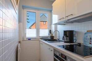 A compact kitchen with white cabinetry, a built-in oven, a coffee maker, and a window with a view of a house. - 1 "Seestern" in Haus Colonia (ID 292) (St. Peter-Bad)