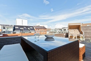 Eine Dachterrasse mit Glastisch, Korbmöbeln und Blick auf die umliegenden Gebäude.