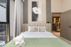 Ein Hotelzimmer mit einem großen Bett, einem grünen Teppich mit Struktur, einem Blumenbild und Blick auf die Stadt durch das Fenster.