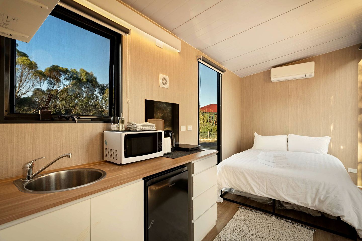 Un dormitorio moderno con cama, microondas integrado y ventana con vista a árboles.