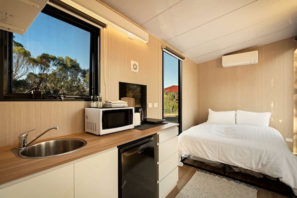 Un dormitorio moderno con cama, microondas integrado y ventana con vista a árboles.