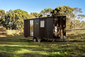 A small, dark-colored mobile home with a sliding door, a water heater, and propane tanks on a trailer.